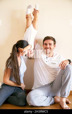 Mama und Papa nehmen das kleine Mädchen an den Beinen auf, während sie im Zimmer auf dem Boden sitzen Stockfoto