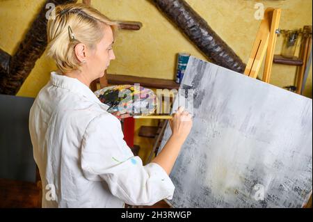 Happy Woman malt mit einem dünnen Pinsel auf Leinwand. Leinwand steht auf der Staffelei. Künstler zeichnet an der Staffelei. Seitenansicht der weiblichen Malerei p Stockfoto