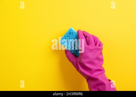 Eine Hand in einem rosa Gummihandschuh hält einen blauen Waschschwamm auf gelbem Hintergrund Stockfoto