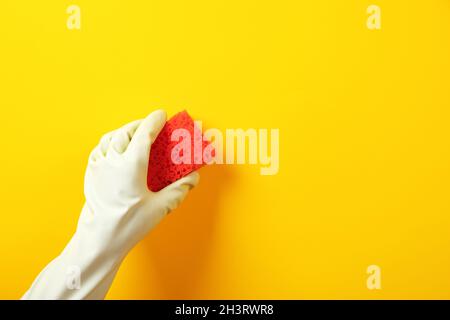 Eine Hand in einem grünen Gummihandschuh hält einen roten Waschschwamm über gelbem Hintergrund Stockfoto
