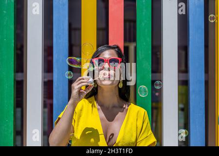 Vorderansicht einer Latina-Frau, die an einem sonnigen Tag Seifenblasen in einem gelben Kleid und Sonnenbrillen vor einem Hintergrund von bunten Bars bläst. Stockfoto