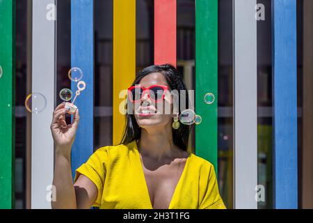 Vorderansicht einer Latina-Frau mit Bubbles Zauberstab in einem gelben Kleid und Sonnenbrille vor einem Hintergrund von bunten Bars an einem sonnigen Tag. Stockfoto