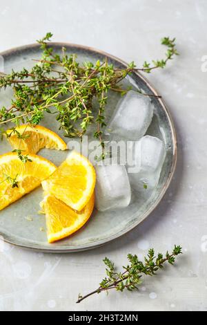 Orangenscheiben, Thymianzweige und Eiswürfel auf dem Teller, Zutaten für einen sommerlichen erfrischenden Cocktail oder kalten Tee Stockfoto