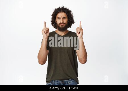 Skeptischer Mann aus der Mitte des osterfestes zeigt die Finger nach oben, runzelt die Stirn und sieht enttäuscht aus, drückt Abneigung aus, steht im T-Shirt auf weißem Hintergrund Stockfoto