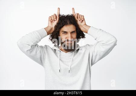 Witziger und hübscher Mann aus der Mitte des ostermondes, der mit einer Geste der Hörner und Stierfingern auf dem Kopf auf weißem Hintergrund stand Stockfoto