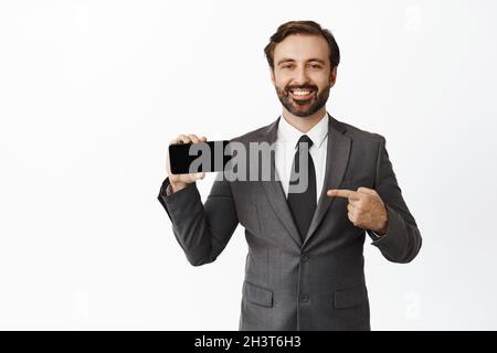 Selbstbewusster Geschäftsmann zeigt mit dem Finger auf den horizontalen Smartphone-Bildschirm und lächelt, während er die Website, die Anwendung und den weißen Hintergrund des Unternehmens vorführt Stockfoto