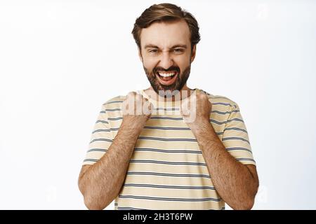 Enthusiastischer bärtiger Mann klatscht mit den Fäusten, lacht und lächelt zufrieden, sieht ermutigt und selbstbewusst aus und steht auf weißem Hintergrund Stockfoto