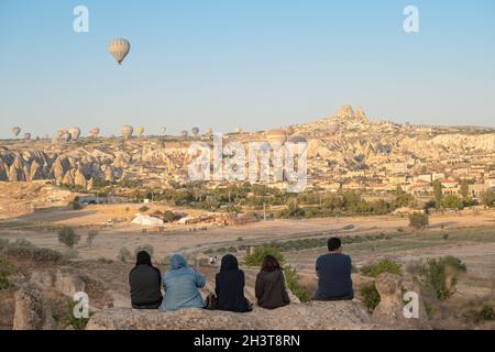 GOREME, TÜRKEI - 5. AUGUST 2021: Morgens beobachten die Menschen auf Felsen sitzend, wie Heißluftballons die Touristen mit uch über das Kappadokien-Tal fliegen Stockfoto