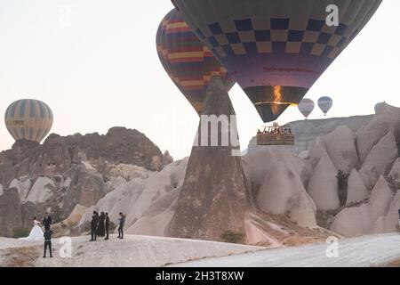 GOREME, TÜRKEI - 5. AUGUST 2021: Menschen feiern eine Hochzeit am Morgen neben den Feen Schornstein Höhlenhäuser, während Heißluftballons mit Touristen Stockfoto