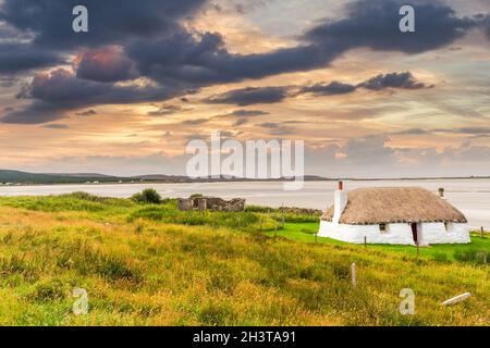 Traditionell gebaute weiße Hütte mit Strohdach, neben der türkisfarbenen Bucht, mit stürmischen bewölkten dunklen Himmel über.Insel North Uist, Schottland Stockfoto