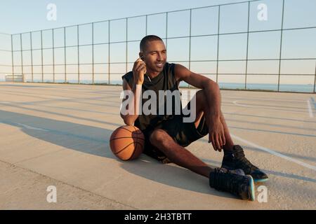 Junger lächelnder afrikanischer Sportler, der auf dem Sportplatz am Mobiltelefon Musik über Kopfhörer hört. Schwarzer Mann in Sportswear und Sneakers. Städtischer Korb Stockfoto