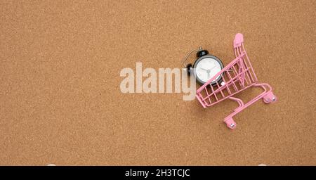 Pinker Einkaufswagen und schwarzer runder Wecker innen auf braunem Hintergrund Stockfoto