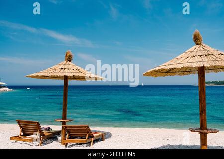 Zwei Holzliegen auf dem weißen Sand, an einem paradiesischen Strand in der Nähe der Stadt Primosten in Kroatien. Stroh-Sonnenschirm. Blauer Himmel Stockfoto