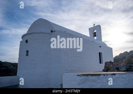 Serifos Island, Griechenland. Kleine alte Kapelle, weiße Wände und Glockenturm, orthodoxe griechische Kirche auf dem felsigen Hügel, blauer wolkiger Himmel Hintergrund. Stockfoto