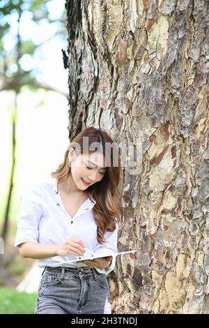 Lächelnde junge Frau, die sich im Park gegen den Baumstamm lehnt und ein digitales Tablet benutzt. Stockfoto