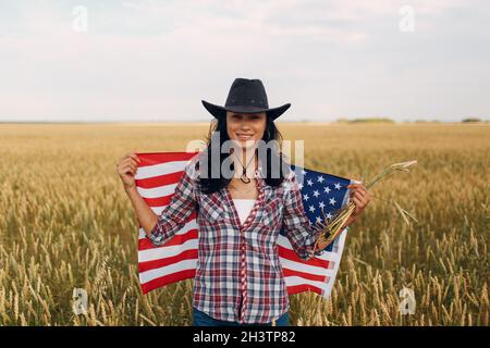 Farmerin trägt Cowboyhut, kariertes Hemd und Jeans mit amerikanischer Flagge auf dem Weizenfeld Stockfoto