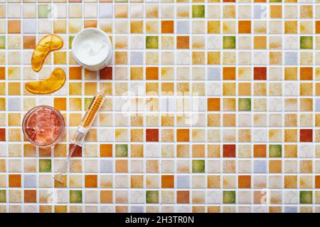 Gläser mit Creme und Gel für die Gesichts- und Körperpflege, goldene Augenflecken und Goldserum in einem Spritzenröhrchen auf dem Hintergrund eines Kachelmosaiks. Tren Stockfoto