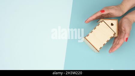 Weibliche Hände gefaltet zueinander über einem hölzernen Miniatur-Modellhaus auf blauem Hintergrund. Immobilienversicherungskonzept, envir Stockfoto