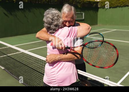 Ältere kaukasische Paare spielen zusammen Tennis auf dem Platz umarmt Stockfoto