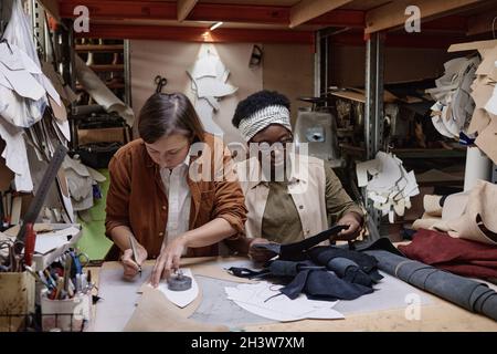 Zwei Schneiderinnen nähen am Tisch Kleidung zusammen, eine Frau zeichnet das Muster und ihre Kollegin schneidet den Stoff in der Werkstatt Stockfoto