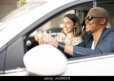Junges glückliches multirassisches Paar, das zusammen am Auto fährt. Ein lateinischer Geschäftsmann auf dem Fahrersitz mit seiner kaukasischen Freundin oder Ehefrau als Pessanger Stockfoto