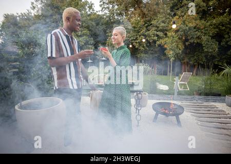 Multirassisches Paar, das abends beim Abendessen in ihrem Hinterhof zusammenhing. Stehen mit Getränken. Konzept der Beziehung. Schwarzer Mann und Stockfoto