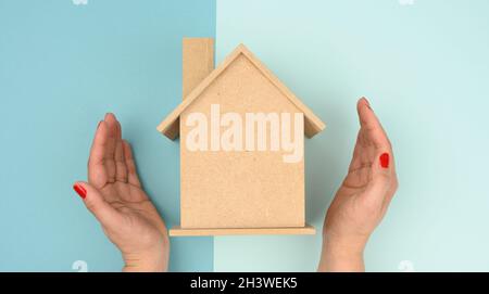 Weibliche Hände gefaltet zueinander über einem hölzernen Miniatur-Modellhaus auf blauem Hintergrund. Konzept der Immobilienversicherung Stockfoto