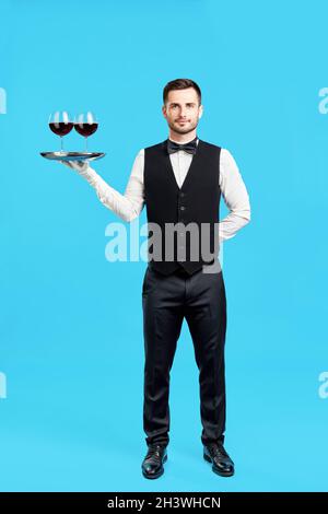 In voller Länge Porträt eines eleganten männlichen Kellners mit Gläsern Wein auf blauem Hintergrund Stockfoto