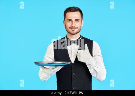 Eleganter junger Kellner mit leerem silbernen Tablett und einem Schild mit Daumen nach oben auf blauem Hintergrund Stockfoto