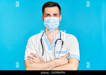 Selbstbewusster Arzt mit medizinischer Maske und gekreuzten Armen auf blauem Hintergrund Stockfoto