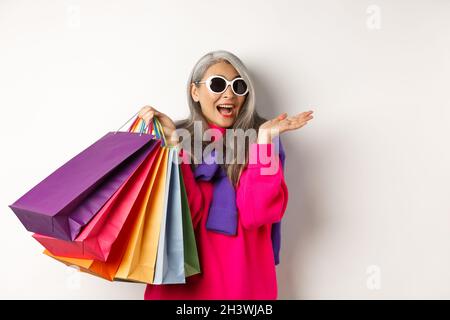 Stilvolle asiatische Senior-Frau mit Sonnenbrille, die im Weihnachtsverkauf einkaufen geht, Papiertaschen hält und lächelt, über weißem Bac steht Stockfoto