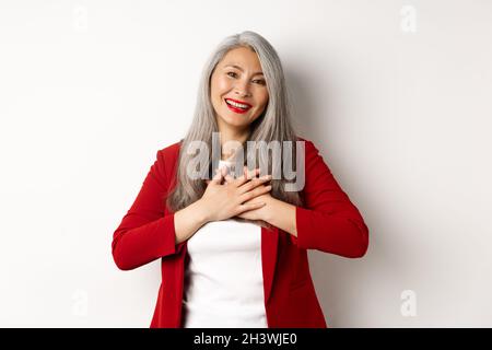Geschäftskonzept. Reife asiatische Frau mit roten Lippen und Blazer, die Hände am Herzen hält und dankbar lächelt und dankbar anschaut Stockfoto