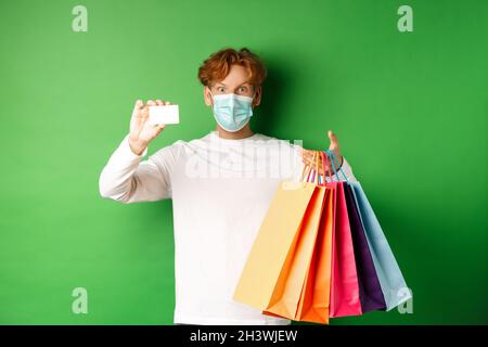 Hübscher junger Mann in medizinischer Maske, zeigt Plastikkreditkarte und Einkaufstaschen mit Artikeln, die mit Rabatt gekauft wurden, grüner Rücken Stockfoto