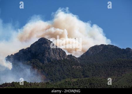 Der Wald an den Hängen eines steilen, felsigen Hügels brennt. Rauch steigt in den Himmel. Wildfire-Hubschrauber am Himmel. Stockfoto