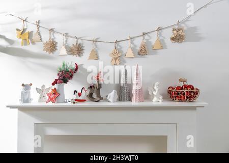 Dekoration des Innenraums der Wohnung zu Weihnachten. Eine Girlande aus geschnitzten Holzfiguren. Kerzen und Keramikfiguren auf einem künstlichen Kamin. Kopie en Stockfoto