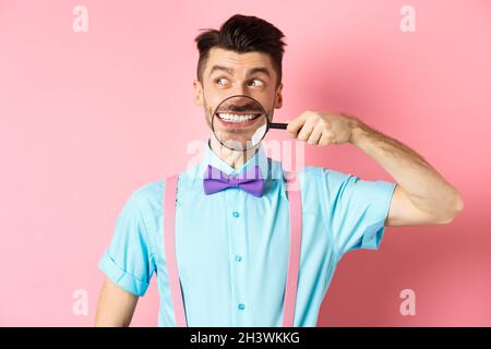 Lustiger Mann in Fliege lächelnd, zeigt Zähne mit Lupe und Blick neben Logo, stand über rosa Hintergrund Stockfoto