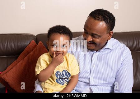 Lachendes Porträt eines 2-jährigen Kleinkindes mit seinem Vater, der zu Hause auf der Couch sitzt Stockfoto