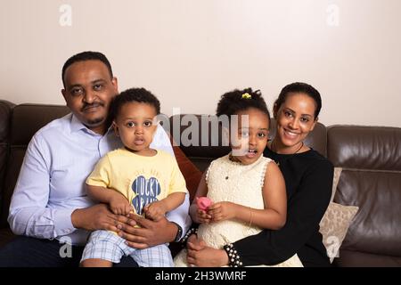 Familie zu Hause, sitzend auf der Couch, 5-jähriges Mädchen, 2-jähriger Bruder, Mutter und Vater, alle schauend auf die Kamera Stockfoto