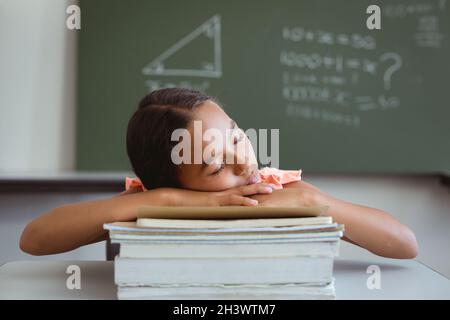 Müde gemischtes Schulmädchen im Klassenzimmer sitzt am Schreibtisch und schläft auf einem Stapel Bücher Stockfoto