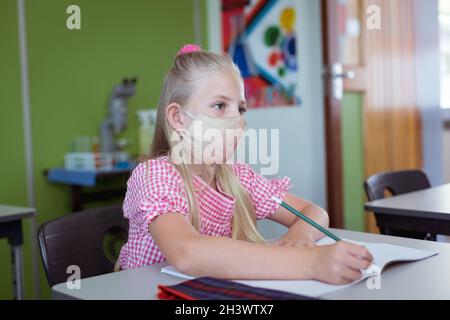 Kaukasisches Schulmädchen, das während des Unterrichts am Schreibtisch im Klassenzimmer mit Gesichtsmaske sitzt Stockfoto