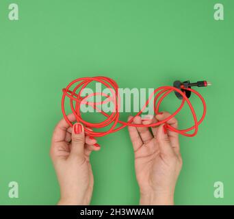 Die weibliche Hand hält verdrilltes Kabel zum Laden von mobilen Geräten in einer roten Textilverpackung auf grünem Hintergrund Stockfoto
