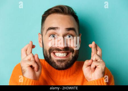 Kopfschuss eines hoffnungsvollen bärtigen Mannes, der einen Wunsch macht, lächelt und die Daumen drückt, um Glück zu haben, und steht über hellblauem Rücken Stockfoto
