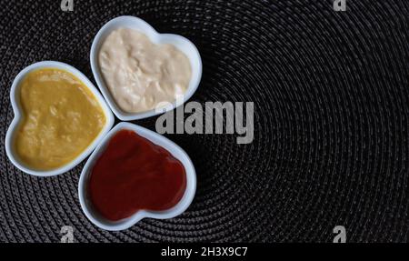 Kleine herzförmige Keramikgläser, gefüllt mit Senf, Ketchup und Mayonnaise, isoliert auf einer Seite, sodass Platz für Kopien bleibt. Stockfoto