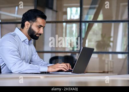 Seriöser ethnischer lateinindischer Geschäftsmann, der in einem modernen Büro mit einem Laptop arbeitet. Stockfoto