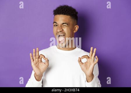 Frech afroamerikanischen Mann winken, stellen Sie sicher, dass Sie in etwas, zeigt okay Zeichen in Zustimmung, stehend zufrieden über lila Rücken Stockfoto