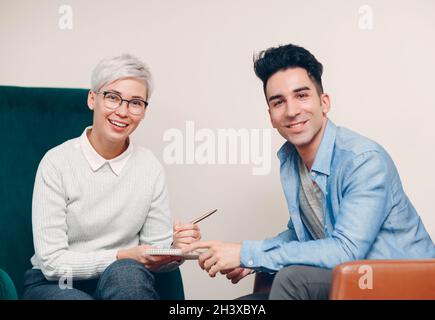 Frau mit Stift und Notizbuch in der Hand schreiben und Mann sitzt auf einem Stuhl zusammen. Journalismus-, Interview- und Personalbefragung c Stockfoto