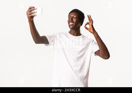 Zufriedener, lächelnder afroamerikanischer junger Mann in Beanie, Selfie oder Videoanruf per Smartphone, mit einer Geste in Ordnung auf sel Stockfoto