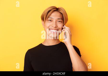Nahaufnahme von schönen stilvollen koreanischen Kerl lächelnd und im Gespräch auf Handy, über gelben Hintergrund stehen Stockfoto