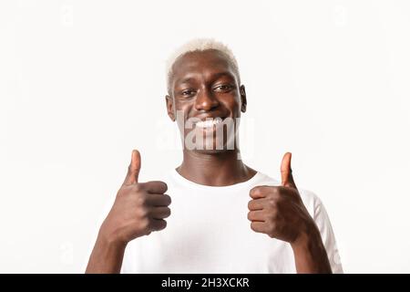 Nahaufnahme von zufrieden gut aussehende afroamerikanische blonde Kerl lächelnd, zeigt Daumen-up in Zustimmung, wie etwas, stehend weiß b Stockfoto