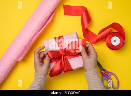 Weibliche Hände binden ein Seidenband auf einer umschlossenen Schachtel aus rosa Papier. Stockfoto